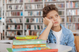 Sad young boy looking away, sitting alone at the library in front of all his books